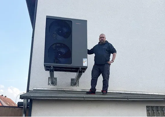 Eine an der Hauswand über einer Garage montierte Wärmepumpe der Firma Waermequelle, daneben stehend ein Mitarbeiter des Teams.