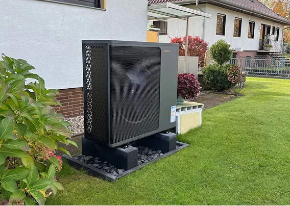 Eine Wärmepumpe der Firma Waermequelle im Garten vor einem Haus mit Rasen.
