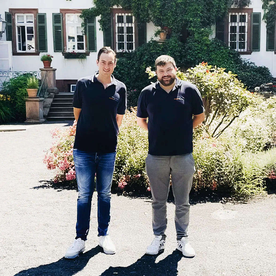 Die Geschäftsführer der Waermequelle GmbH, Youri Kissner und Felix Cvecko, vor einem grünen Busch.