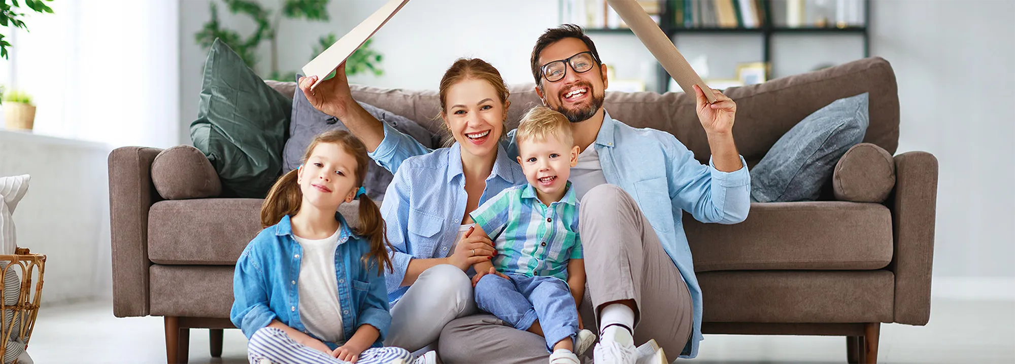 Eine vierköpfige Familie auf dem Boden vor einem Sofa sitzend, während der Vater einen Karton in Form eines Dachs über sie hält.