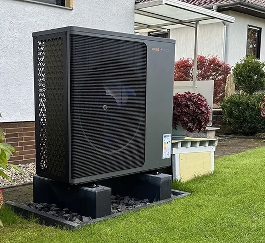 Eine Wärmepumpe der Firma Waermequelle im Garten vor einem Haus mit Rasen.