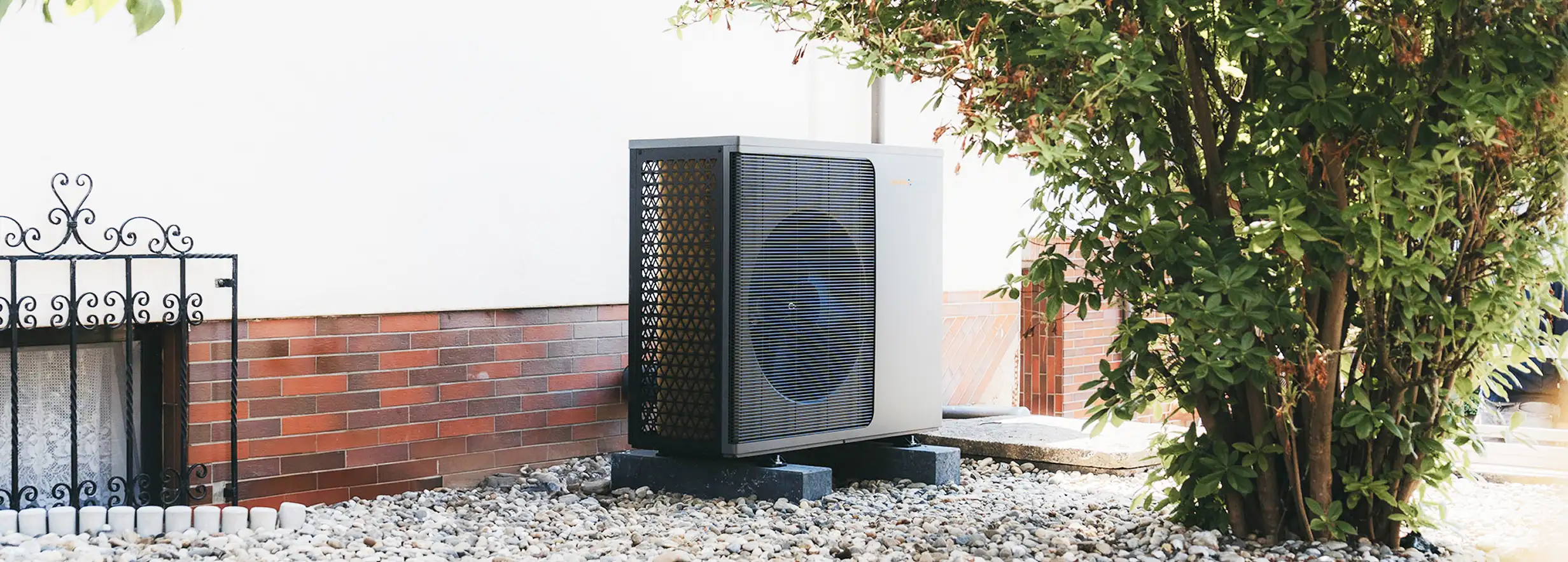 Eine Wärmepumpe der Firma Waermequelle auf einem Kiesuntergrund und neben einem grünen Strauch vor einem Haus.