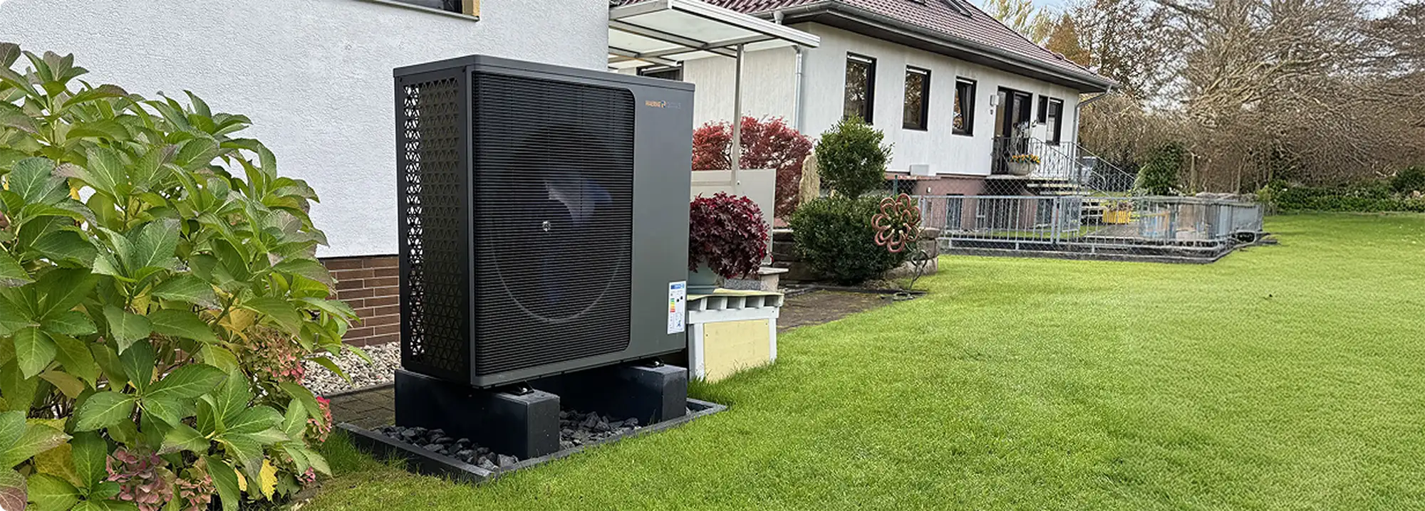 Eine Wärmepumpe der Firma Waermequelle im Garten vor einem Haus mit Rasen.