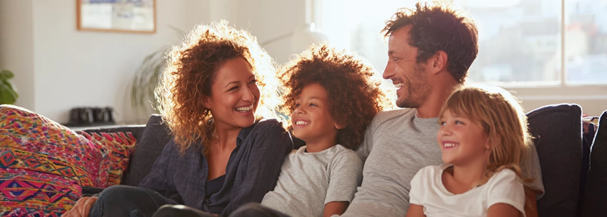 Eine Familie mit Mutter, Vater, Sohn und Tochter, lachend auf einem Sofa nebeneinander sitzend.