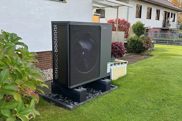Eine Wärmepumpe der Firma Waermequelle im Garten vor einem Haus mit Rasen.