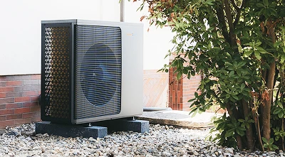 Eine Wärmepumpe der Firma Waermequelle auf einem Kiesuntergrund und neben einem grünen Strauch vor einem Haus.