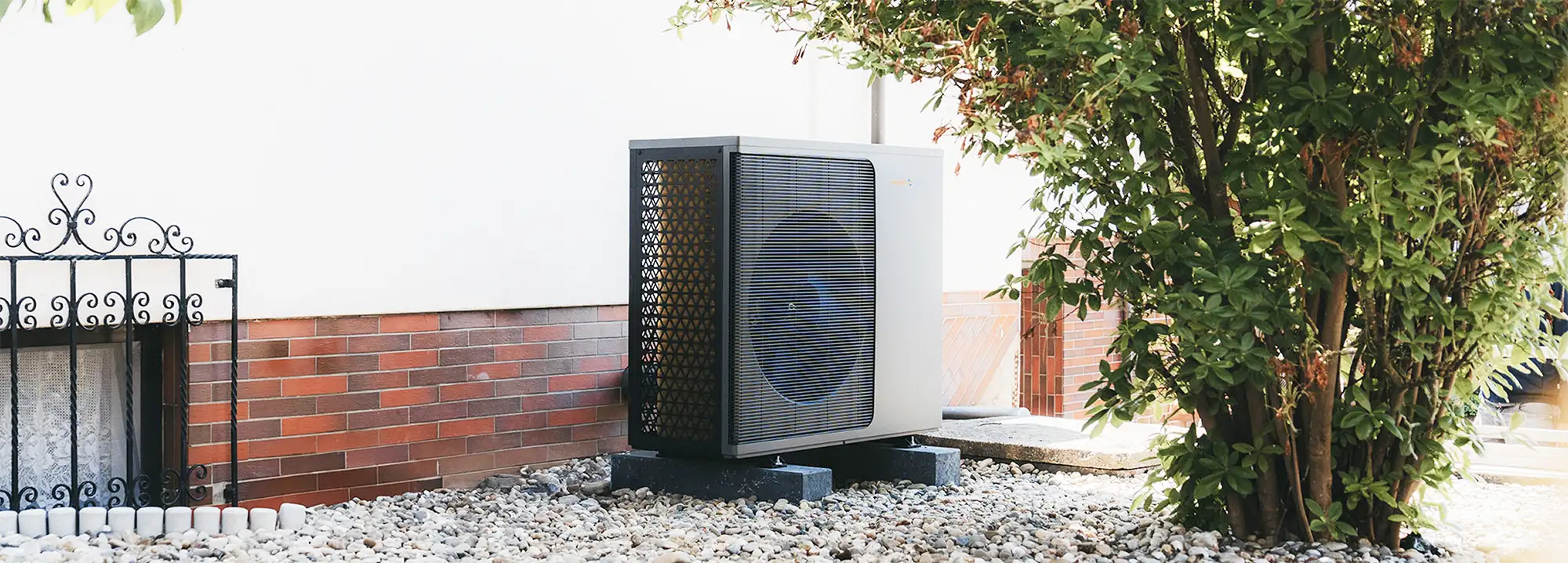 Eine Wärmepumpe der Firma Waermequelle auf einem Kiesuntergrund und neben einem grünen Strauch vor einem Haus.