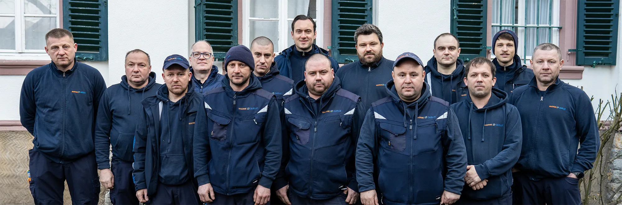 Gruppenbild des Teams der Waermequelle GmbH in dunkelblauer Arbeitskleidung vor einer Hauswand.